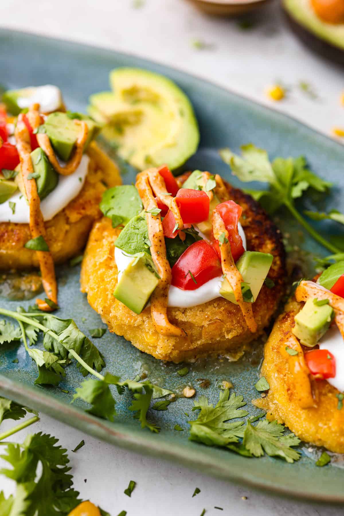 Angle shot of sweet corn tamale cakes plated and topped with fresh tomatoes, avocado, cilantro and sauce.
