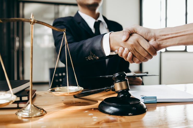 A lawyer shaking hands with a law firm marketing agency representative over a table with a brass balance, a black gavel, three stacked books, and some papers bound with a paperclip.
