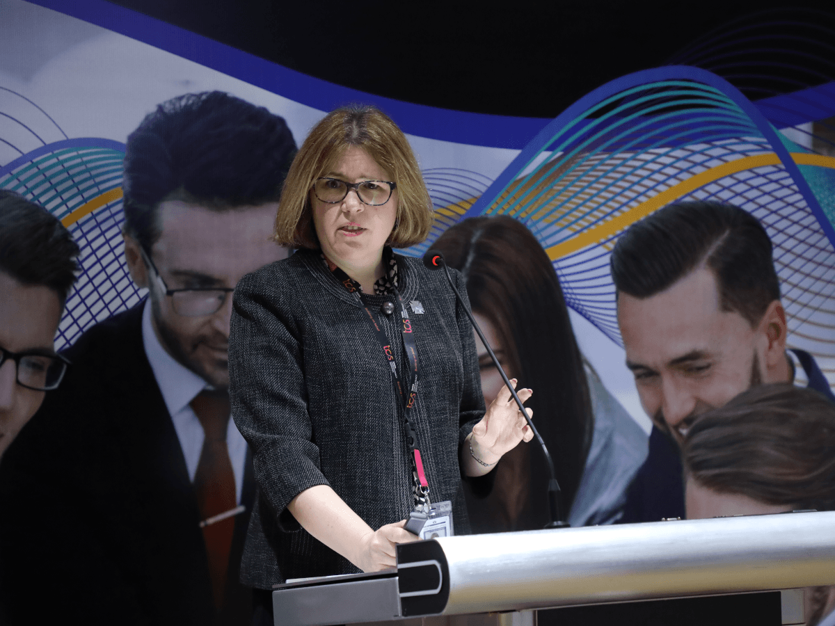 A woman with short hair and glasses stands at a podium giving a talk.