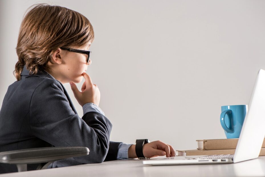 A child in a suit and glasses sits at a desk, contemplating kid business ideas while gazing thoughtfully at a laptop. A blue mug and closed book rest beside him.