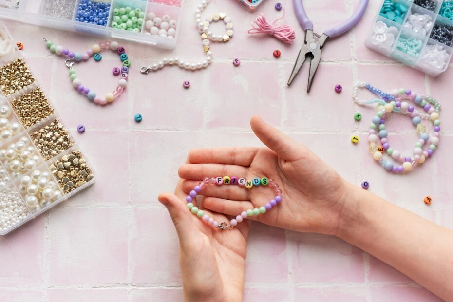 Hands holding a beaded bracelet with the word "FRIENDS" on it, surrounded by various beads, a finished bracelet, pliers, and containers of beads—perfect crafty things to sell at school—on a pink tiled surface.