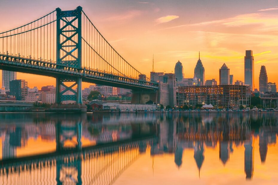 A romantic view of the Philadelphia skyline, water, and bridge at sunset.