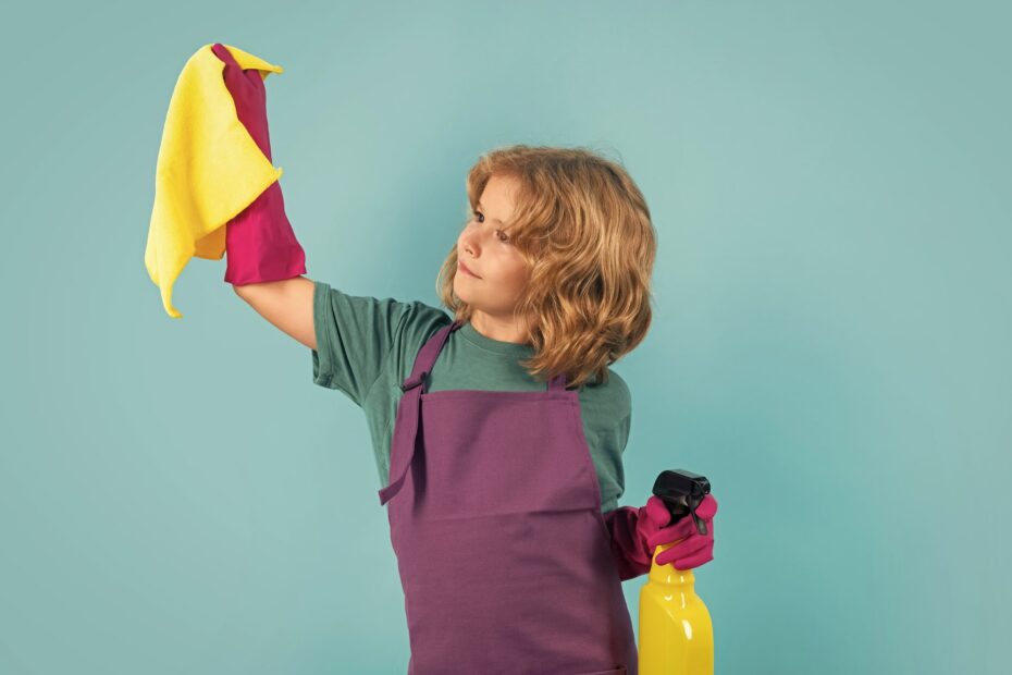 A child wearing an apron and gloves stands against a teal background, holding a yellow cleaning cloth and a spray bottle, ready to do some chores to make money.