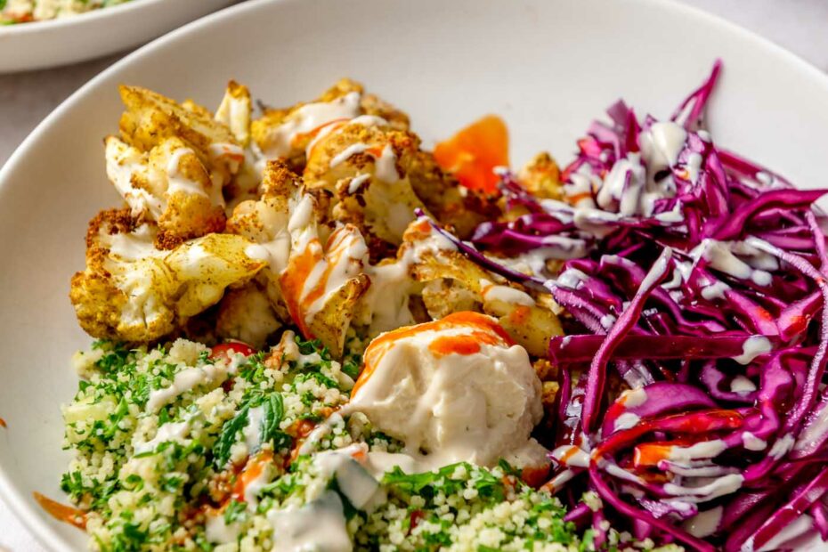 Cauliflower shawarma bowl with roasted spiced cauliflower, shredded cabbage, couscous tabouleh, hummus and tahini sauce and hot sauce drizzled over top.