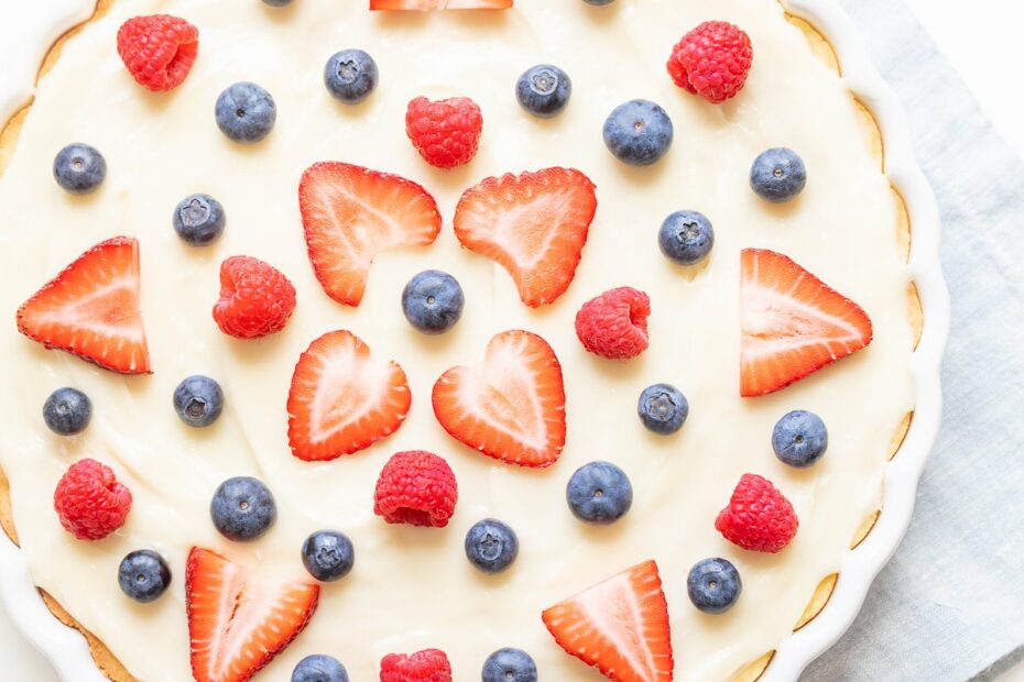 A fresh fruit pizza with cream and a decorative arrangement of strawberries, blueberries, and raspberries, finished with a glaze.
