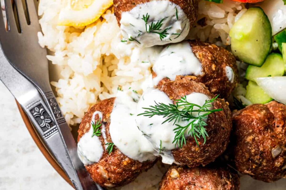Overhead view of baked chicken meatballs topped with garlic dill yogurt sauce served over rice, next to a side of salad on a plate.