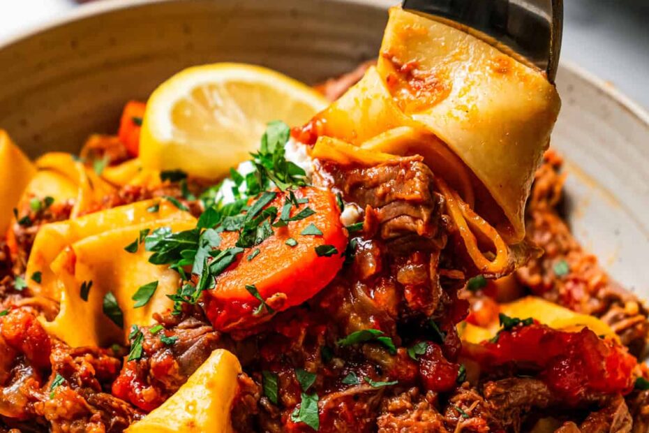 Close up overhead view of beef ragu with pappardelle pasta in a bowl garnished with parsley and a lemon wedge.