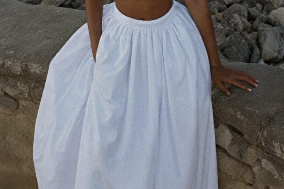 Person in a white sleeveless crop top and long skirt leaning against a stone wall by a rocky shoreline, embodying the effortless elegance of French Riviera travel outfits. The ensemble contrasts with the rugged background, creating a serene and stylish scene.