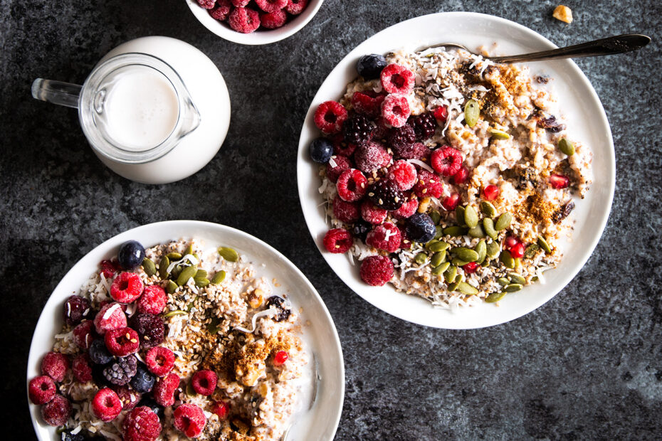 Two bowls of Power Porridge topped with raspberries, pumpkin seeds, coconut, chia seeds, sesame seeds, date sugar, coconut milk, and pomegranate seeds.