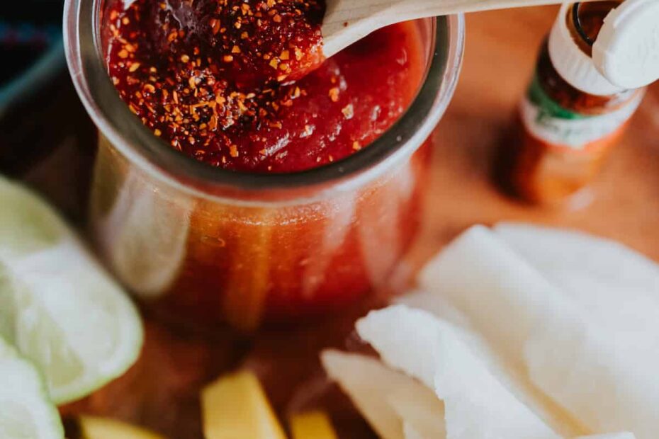Homemade Mexican Chamoy Sauce in a preserve with a wooden spoon and sprinkled with chile lime seasoning and slices of mango and jicama on plate.