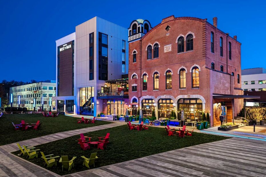 A nighttime exterior view of Hotel West & Main showcasing the dramatic architectural contrast between the preserved 1874 red brick firehouse (right) with its distinctive arched windows and decorative elements, and the modern hotel tower addition (left) with its contemporary facade. The buildings are beautifully lit against a deep blue evening sky. The courtyard features colorful Adirondack chairs in firefighter-inspired red, yellow and green hues arranged on a grassy area with wooden walkways, creating an inviting outdoor social space.