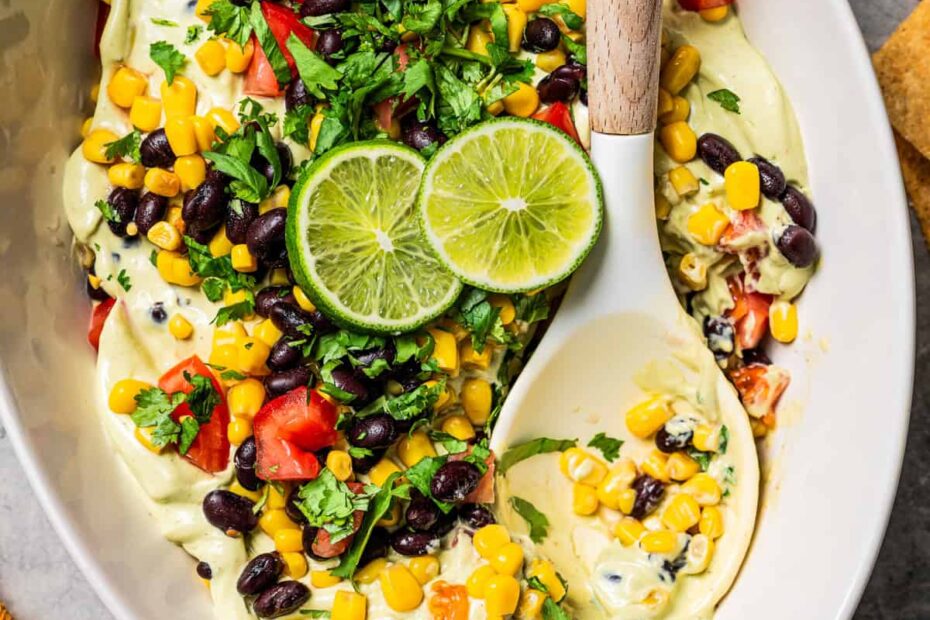 Layered avocado dip topped with lime wedges and fresh cilantro in a serving dish, with a spoon.
