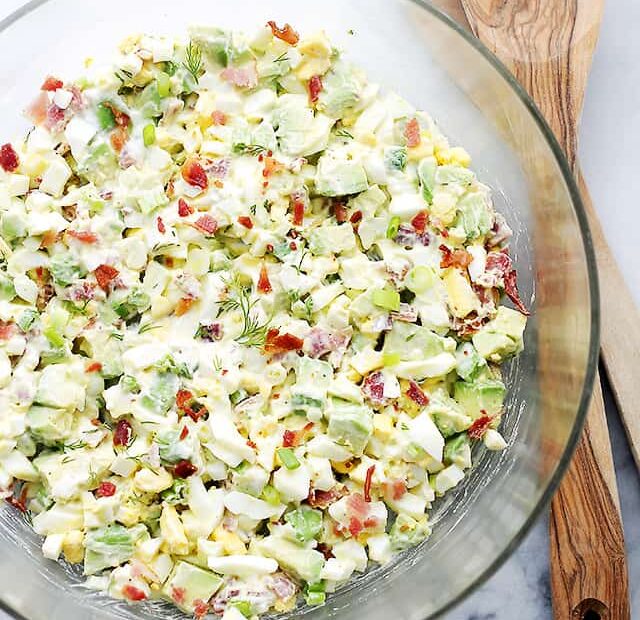 Overhead view of avocado egg salad in a glass bowl with two wooden serving tongs.
