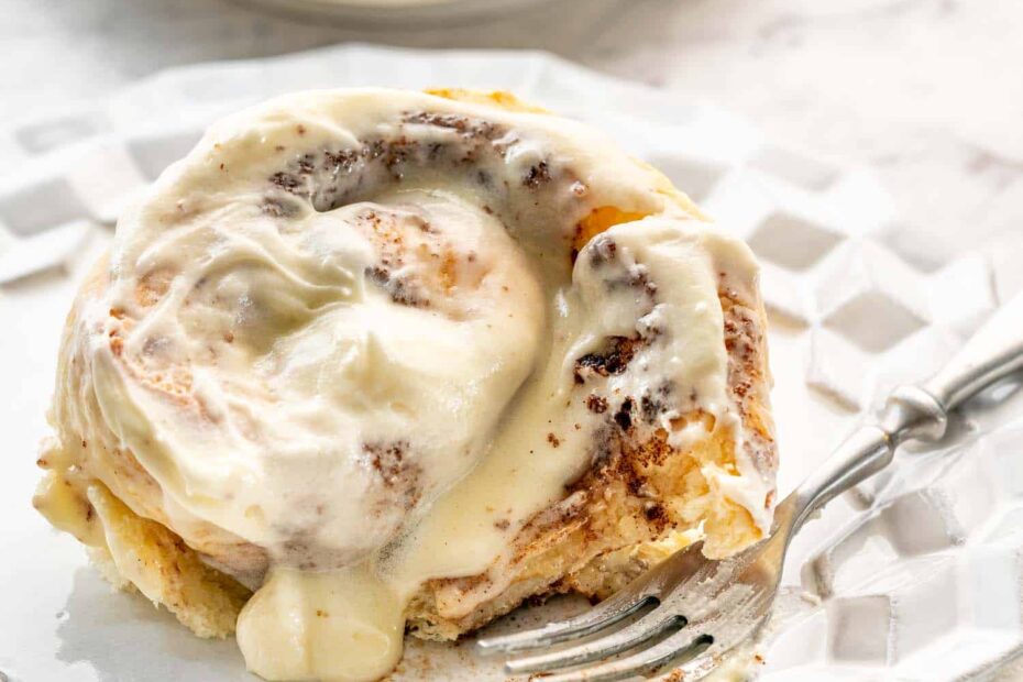 a cinnabon cinnamon roll on a white dessert plate with a cup of coffee in the background.