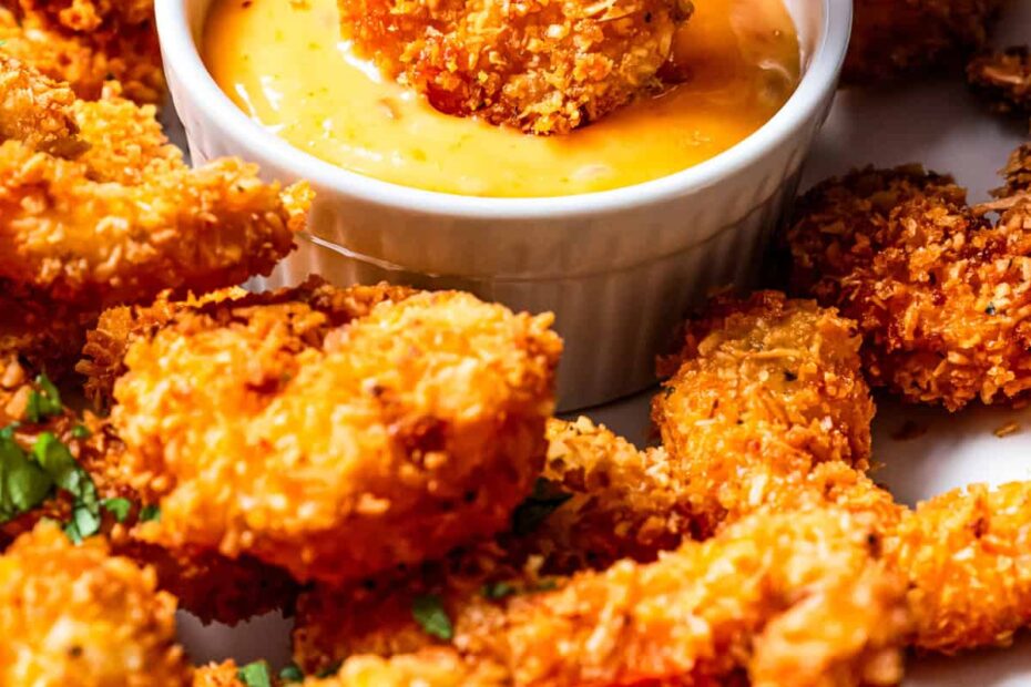 A hand dipping a coconut shrimp into a small bowl of spicy mayo dipping sauce, surrounded by more coconut shrimp on a white plate.