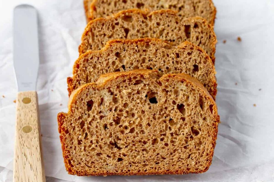 Four slices of banana bread are arranged on parchment paper with a butter knife on a wooden board.