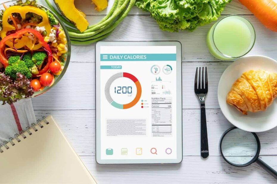 An overhead view of a tablet featuring a daily calorie counting tracker, surrounded by a salad, juices, vegetables, a croissant, a fork, and a notebook.