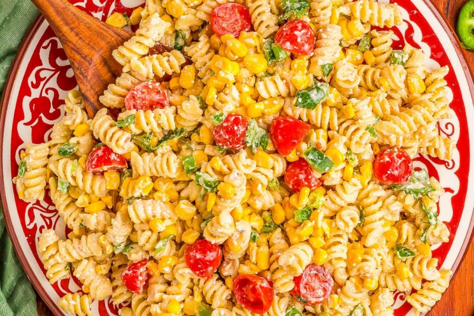 A plate of rotini pasta salad with cherry tomatoes, corn, green onions, cilantro, and a creamy dressing, surrounded by lime wedges, jalape&ntilde;os, tomatoes, and spices on a wooden table.