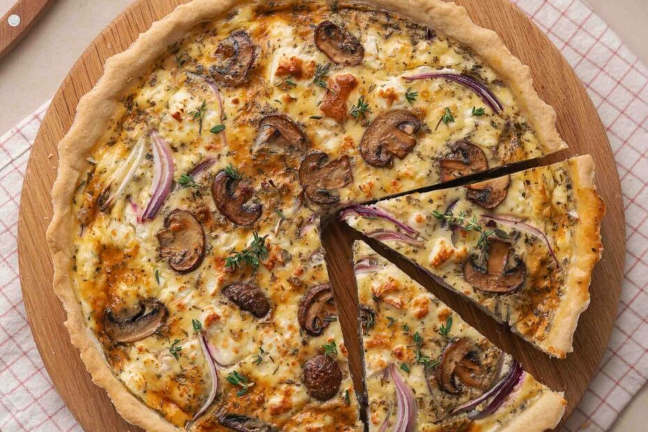 A quiche baked with mushrooms, onions and herbs on a wooden board, with a slice cut out. Nearby are a knife, greens, pepper mill and minced onions on a tea towel.