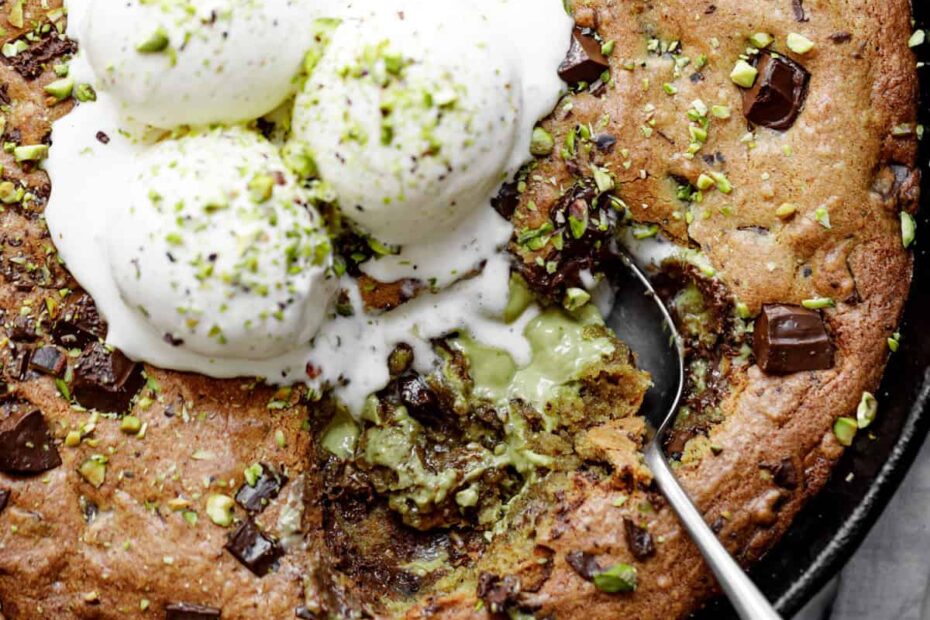 Close-up image of the spoon lifting a portion of pistachio cookie from a skillet, served with vanilla ice cream and topped with chocolate.