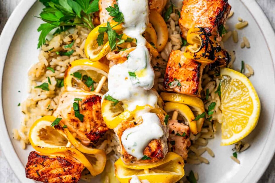 Salmon skewers served over a bed of rice on a plate, topped with garlic sauce and garnished with lemon wedges.