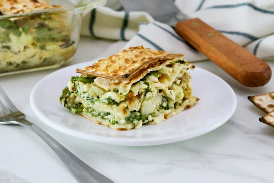 Close up of a slice of spinach matzo pie with layers of spinach, feta cheese, and artichokes, baked to perfection and served on a white plate, fork and towel beside the plate. Spinach matzo mina pie in background.