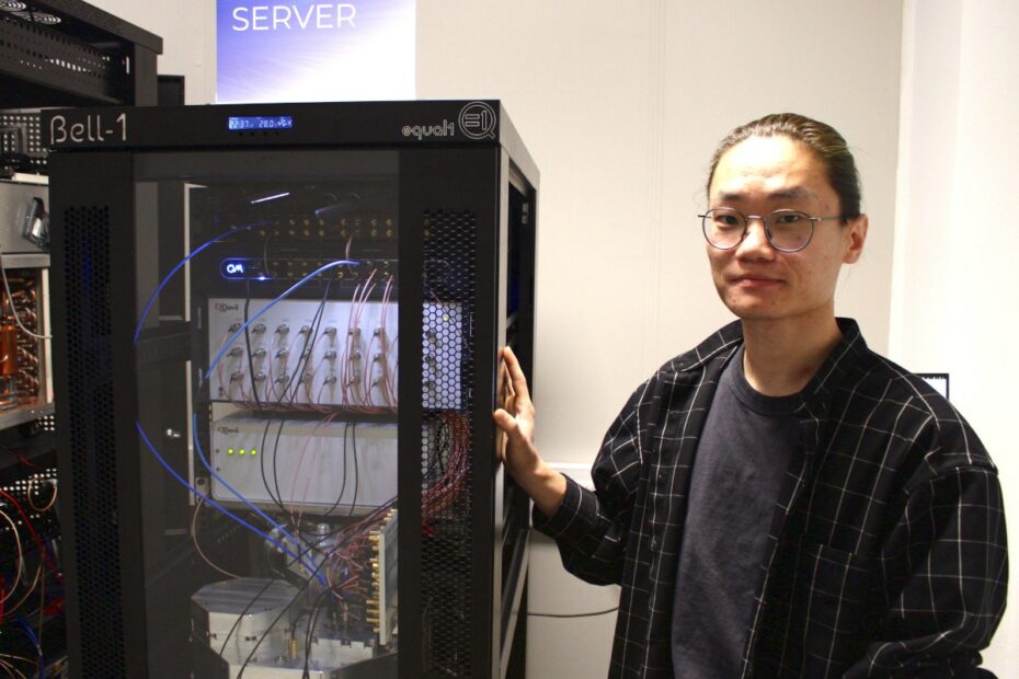 A man wearing glasses and a check shirt over a grey T-shirt stands next to a quantum processing unit.