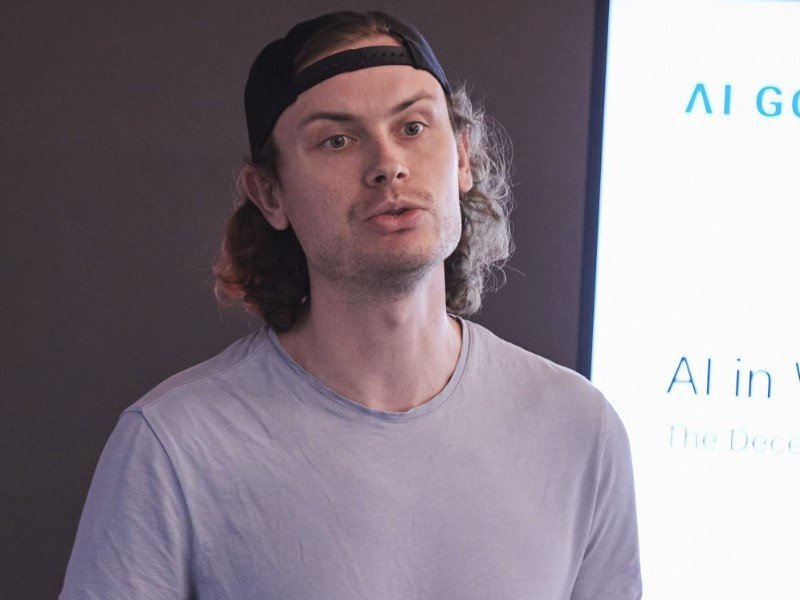 A close-up of Richard Blythman giving a presentation in front of a screen. He wears a cap backwards and a pink t-shirt.