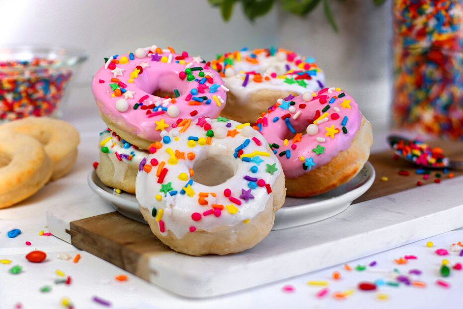 Multiple air fryer donuts with pink and white glazes covered in rainbow sprinkles arranged on a white marble cutting board, with jars of colorful sprinkles visible in the background and plain unglazed donuts to the side.
