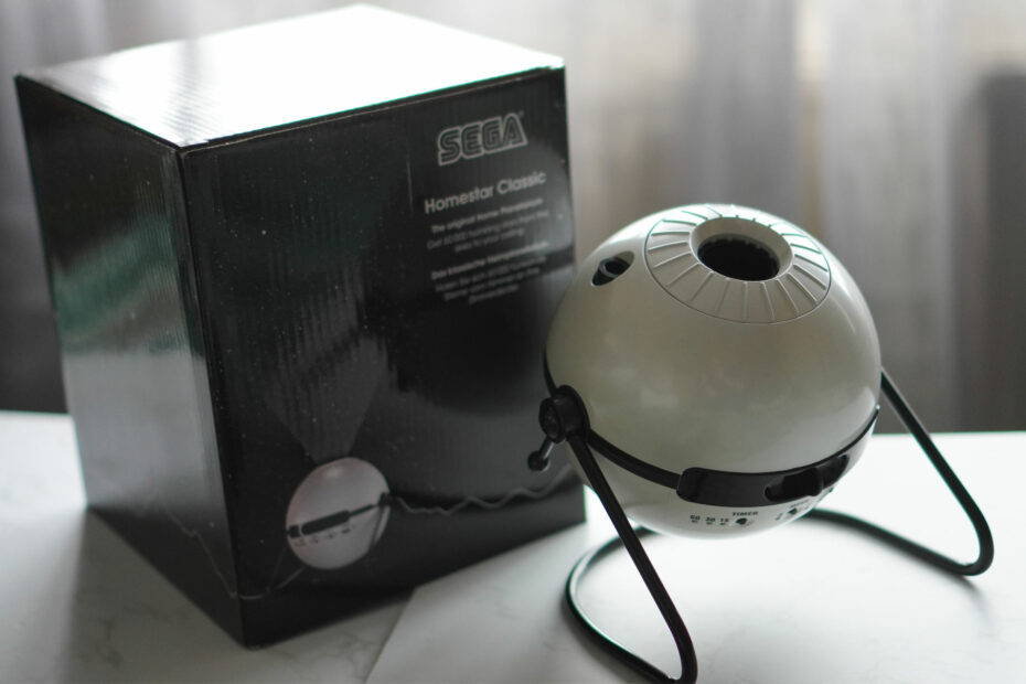 A white, spherical projector is placed on black legs on a white surface next to a black product box with white curtains behind it.