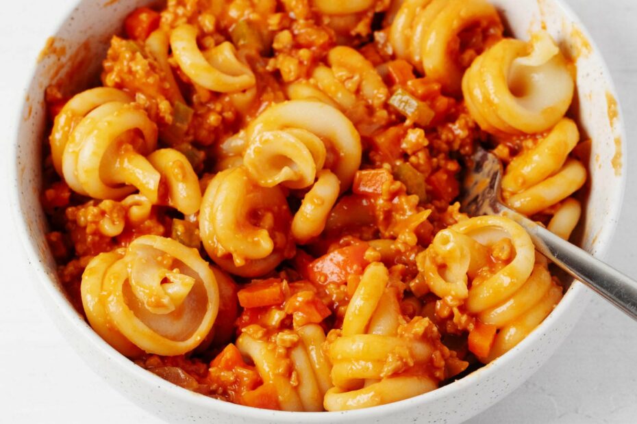A round, white ceramic bowl rests on a white surface. It holds curly pasta shapes with a vegan bolognese sauce.