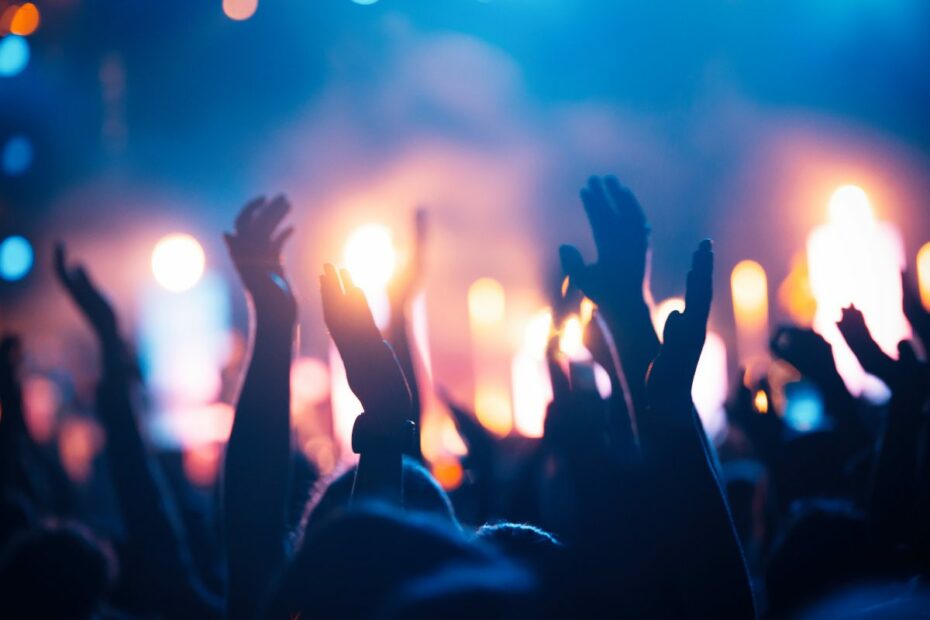 The raised hands of a crowd at a music show at night facing blurred lights of the stage.