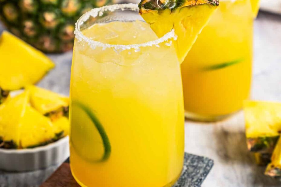 A pineapple margarita in a salt-rimmed glass garnished with a pineapple wedge and lime slice, with a second margarita in the background. Photo by diethood.