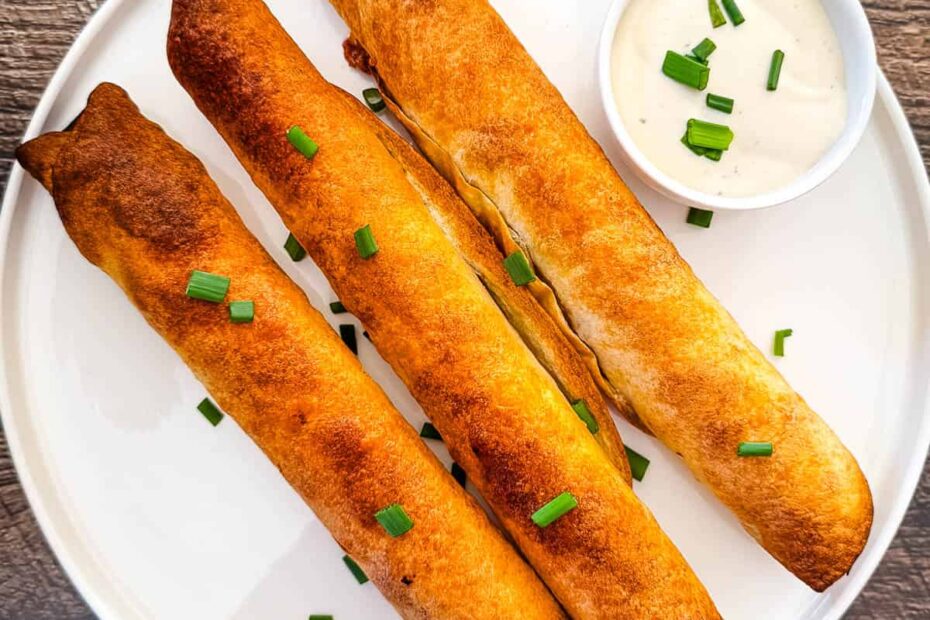 Three golden-brown baked pulled pork taquitos on a white plate, garnished with chopped green onions and served with ranch dipping sauce.