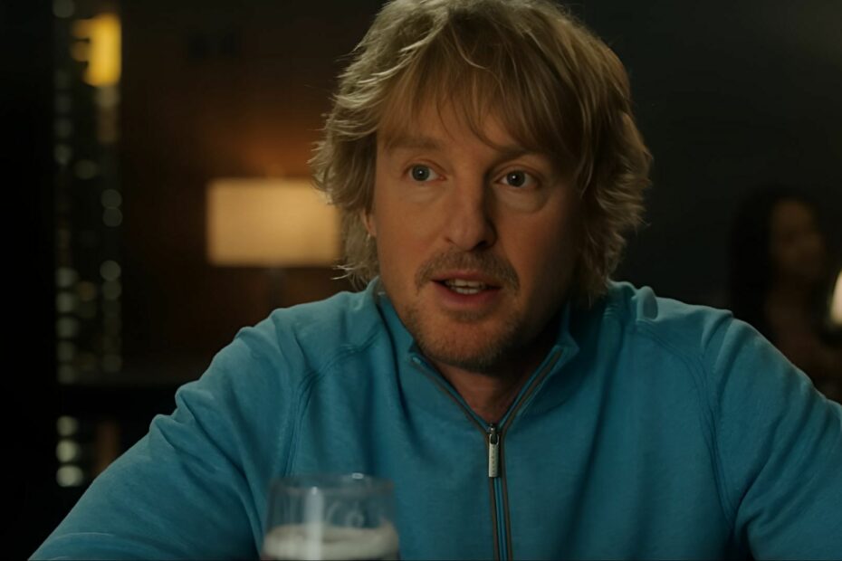 Owen Wilson in a blue pullover, speaking while seated at a bar table with a drink in front of him.