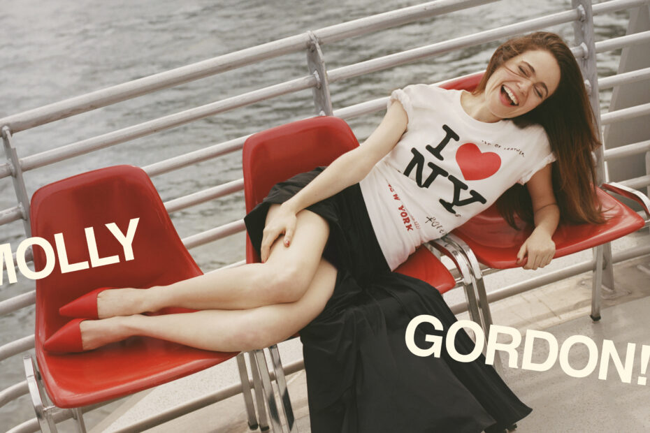 Molly Gordon sitting across three red chairs outside on a ferry boat. She is wearing an "I Love NY" t-shirt with a black skirt and red heels. On the image, it says "Molly Gordon" with an exclamation point.
