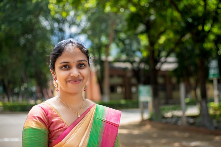 A young woman wears a colourful sari and stands outside smiling at the camera.