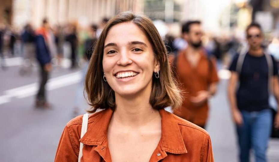 A blonde woman in the middle of the street, smiling and walking around with an orange jacket and a bag.