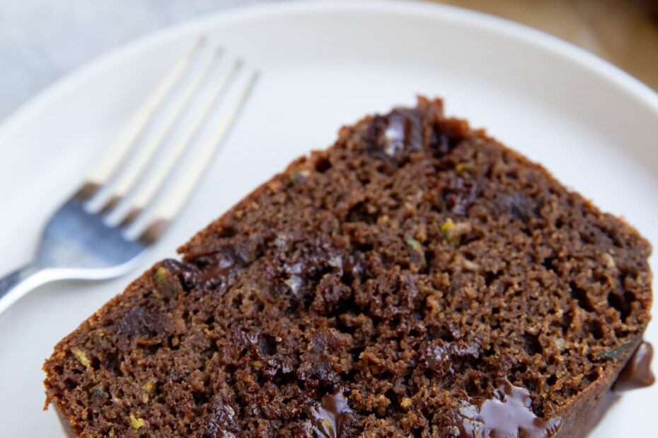 Slice of chocolate zucchini bread on a white plate with the rest of the loaf sliced in the background.
