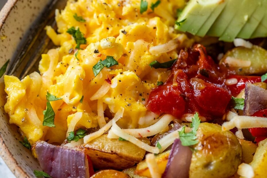 A breakfast bowl of scrambled eggs, roasted potatoes, avocado slices, cheese, salsa, and herbs, with a fork on the side.
