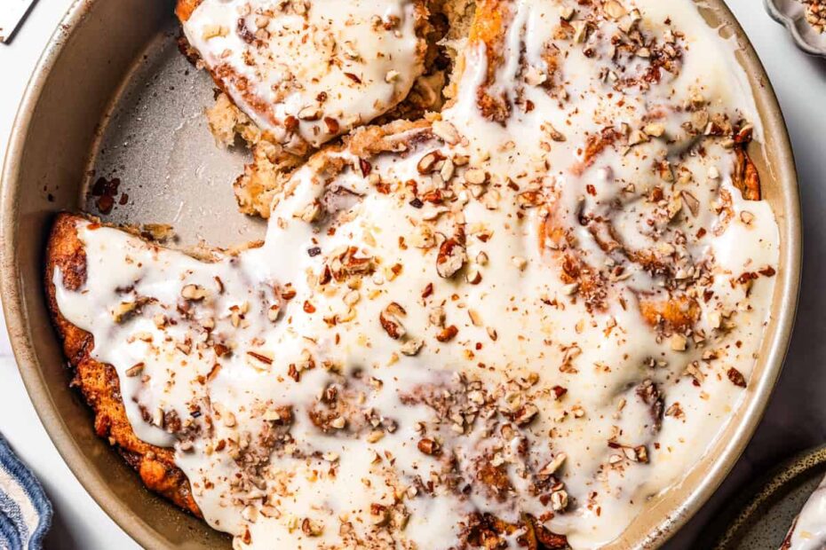 Frosted cinnamon rolls topped with pecans in a round baking pan, with one cinnamon roll missing.