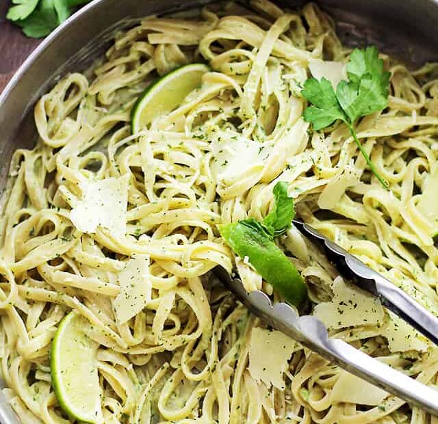 A set of tongs resting in a skillet of avocado pasta garnished with shaved parmesan and basil leaves.