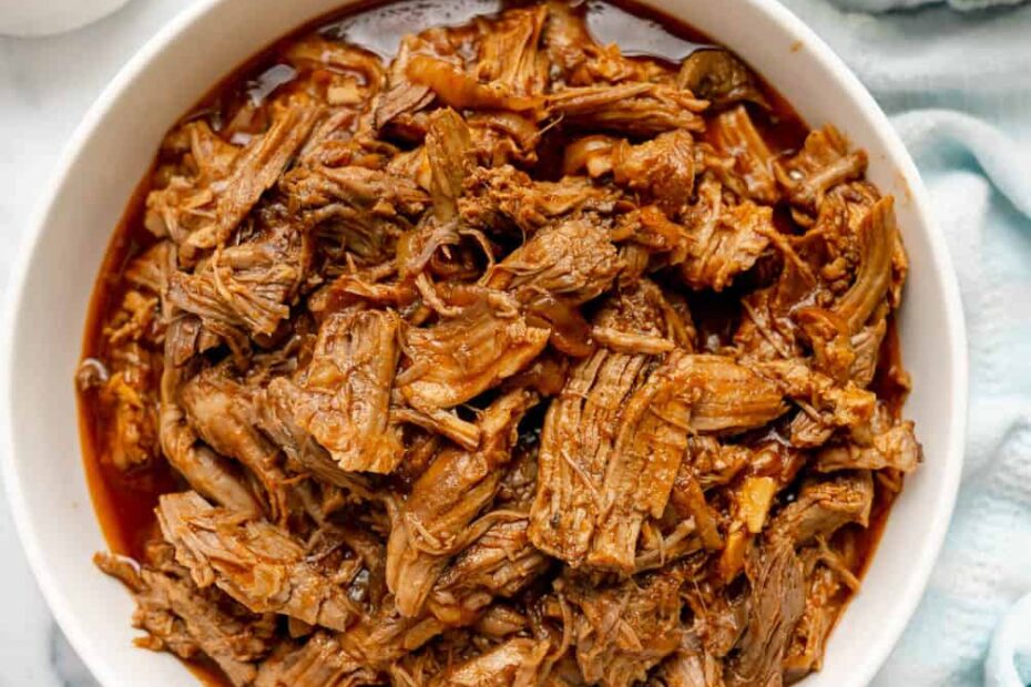 A bowl of shredded barbecue pulled pork sits on a white surface, surrounded by small bowls of pickles, barbecue sauce, and coleslaw.