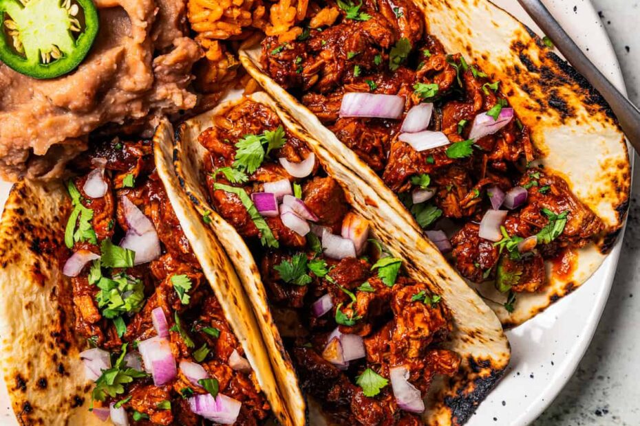 Three chilorio tacos served with a side of seasoned rice and refried beans on a plate. Photo by diethood.