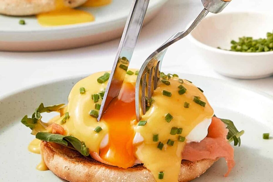 A fork and knife cutting into a poached egg on toasted English muffin with creamy hollandaise sauce and chives — the yolk just starting to spill out.