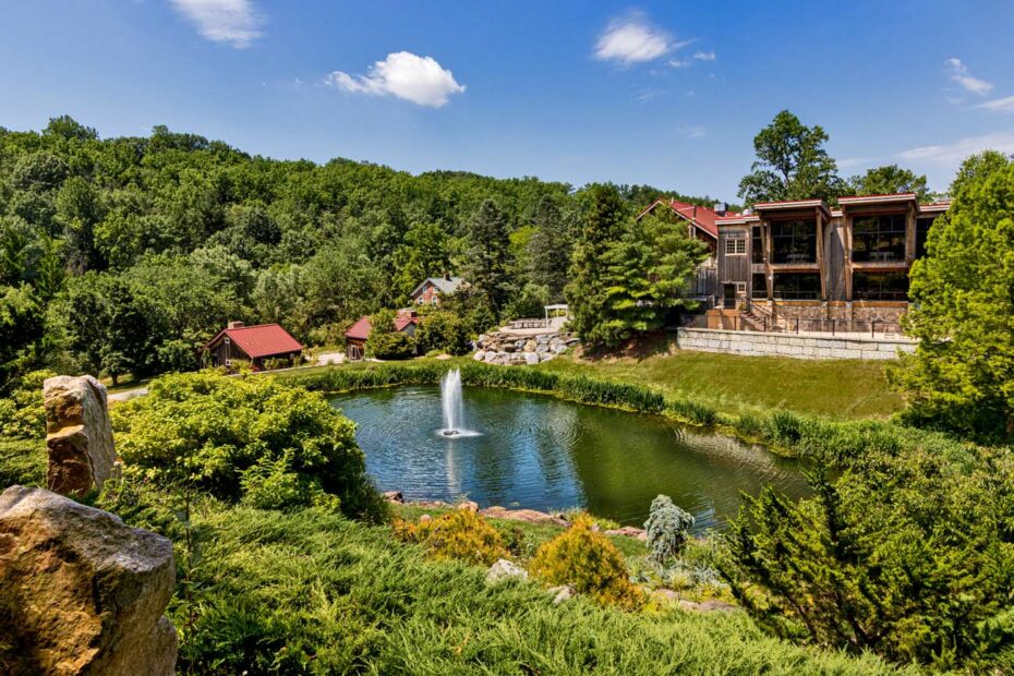 Panoramic view of Glasbern's main pond and buildings. Elevated view showing a large pond with central fountain surrounded by landscaped grounds, with multiple rustic buildings including barns and guest accommodations scattered throughout the wooded property