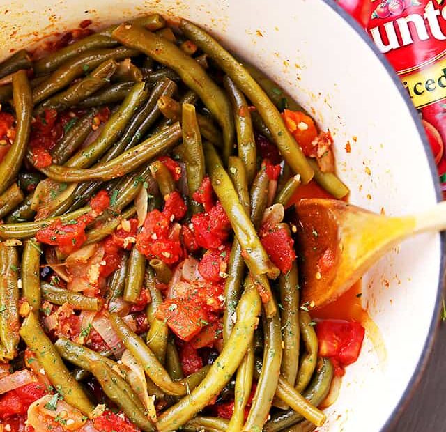 Overhead view of braised green beans and tomatoes in a pot with a wooden spoon.