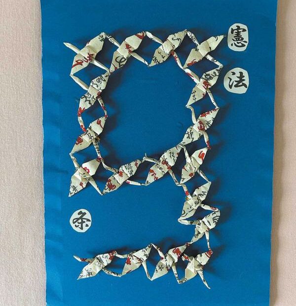 A blue foam board carries the Japanese word "Constitution" at its upper right corner. At the center, 18 hand-folded paper cranes are arranged to form the number "9" — symbolizing Article 9 of the Constitution of Japan, the peace clause. JIANG XUEQING/CHINA DAILY