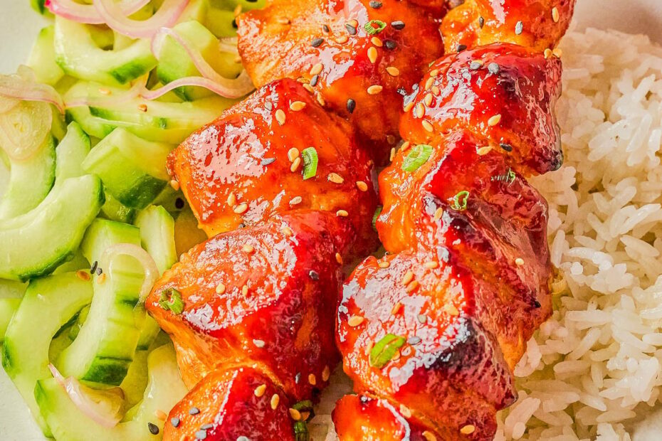 Two skewers of glazed grilled chicken served on white rice with a cucumber salad on a white plate.