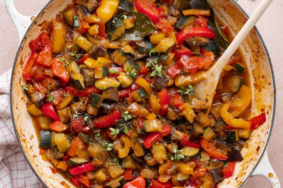 A white pot filled with cooked ratatouille, consisting of chopped zucchini, eggplant, peppers and tomatoes, sits on a checkered napkin with a wooden spoon and fresh herbs nearby.